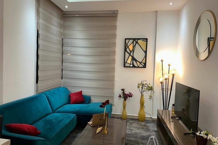 Un salon cosy avec un canapé en velours bleu turquoise, parsemé de coussins rouge. Une lampe sur pied éclaire doucement la pièce, créant une atmosphère invitante. les murs sont décorés de tableau et de miroir. Une table basse assortie avec le meuble Tv, un tapis couvrant tout l'espace ainsi que des vases de couleur et objets décoratifs